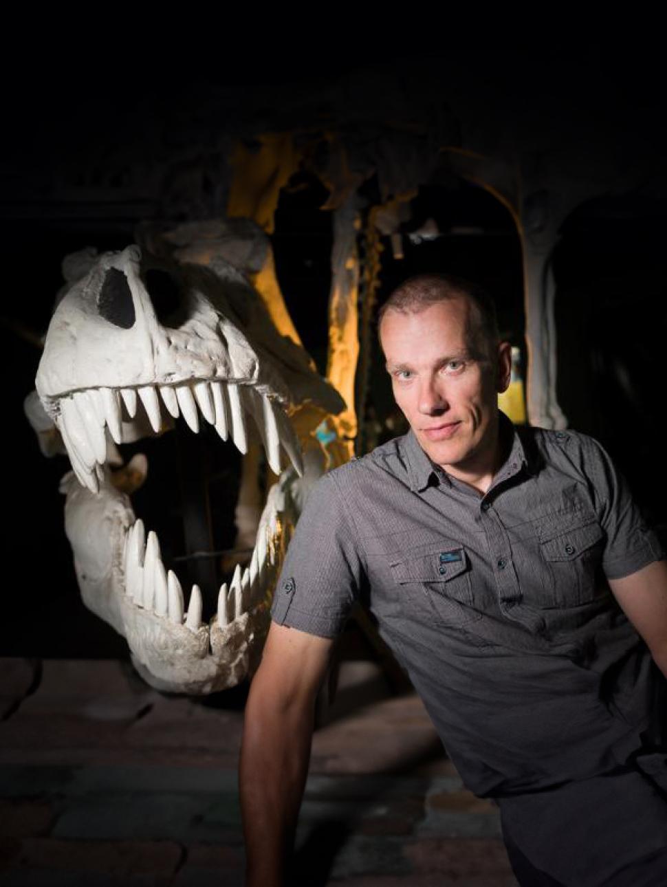 Dr Mikael Trex wears a grey polo shirt and sits in front of a large white cast of a dinosaur skull under cinematic lighting. They stare into the camera with a friendly gaze
