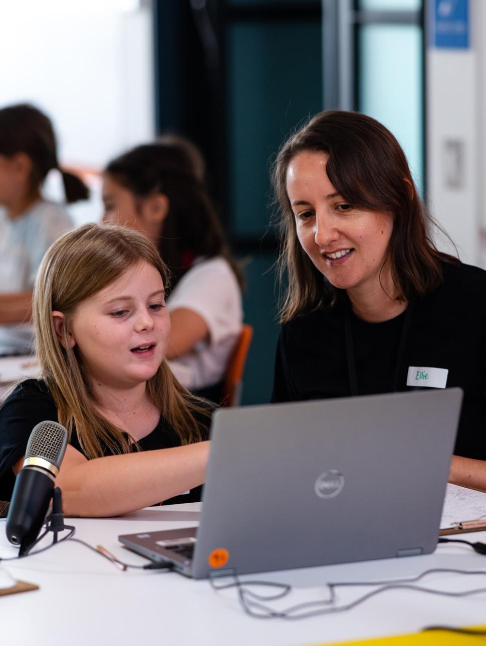 a child sits at a table with laptop and a microphone. an adult sits nearby assisting