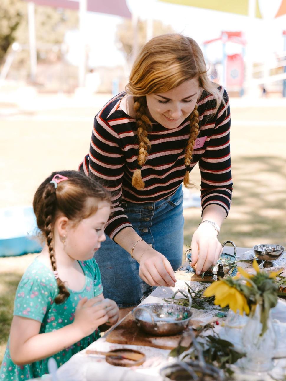 A young child in a blue dress with plaited hair and an adult in a striped shirt with auburn plaits stand over a table filled with messy play objects and tools outside on a sunny day