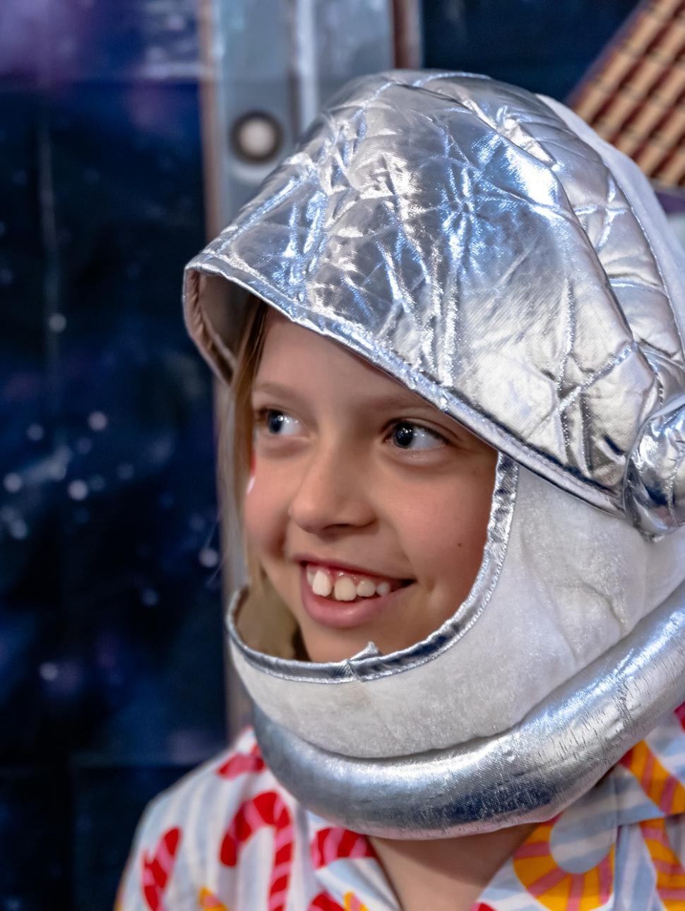 A child wears a dress up space helmet and costume and smiles in front of a space backdrop 