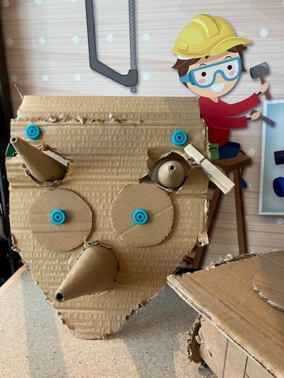 A piece of brown cardboard is fashioned into a haphazardly constructed triceratopcs head with three conical horns and two large round eyes. Small blue screws hold the piece together and a cartoon of a young boy in a yellow builders hat hangs in the background