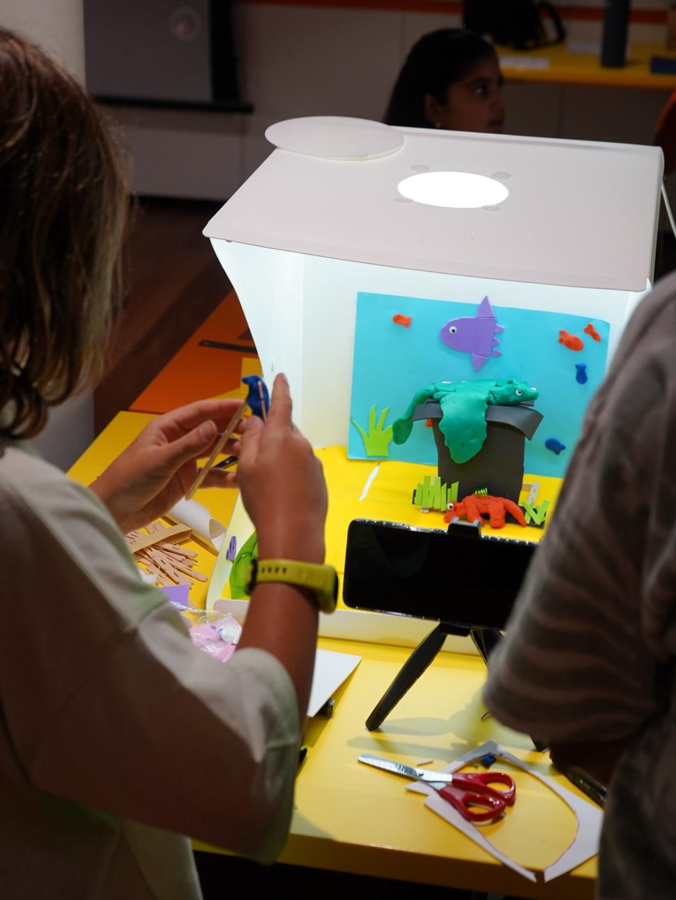 A claymation photography set up on a yellow table. A child fashions a piece of colourful clay to add to an underwater themed clay diorama brightly lit by a white light box and positioned in front of a camera to film.