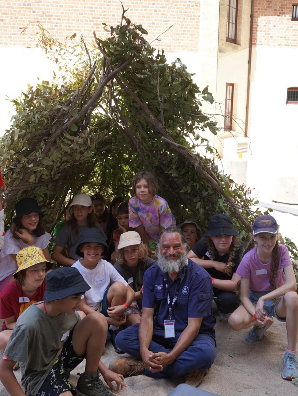 A group of 14 young children in an array of colourful t-shirts, shorts and bucket hats sit proudly behind Derek Nannup who has a long white beard and wears navy blue trousers and a shirt. The group are seated inside, and in front of a large triangular structure made of branches and leaves in a sandpit