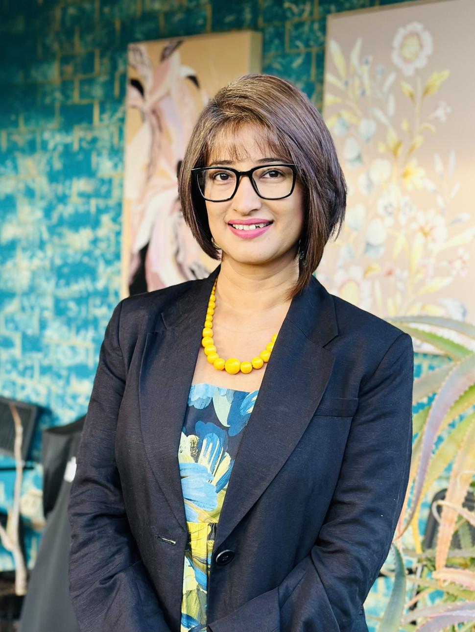 Dr Parwinder Kaur stands against a blue and peach backdrop with flower motifs and a large plant behind her. She has short dark brown hair cut into a bob and wears black rimmed glasses. She is wearing a black suit jacket over a floral printed shirt that matches the backdrop and a chunky yellow necklace. She is looking at the camera a smiling slightly   