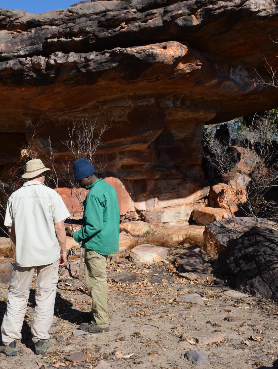 Two men stand at a rock art site, one wearing khaki trousers, a khaki short-sleeved shirt and khaki hat and the other wearing brown trousers, a bright green short and a dark blue beanie. They seem engaged in conversation as they stand underneath a vibrant red orange and black banded rock formation