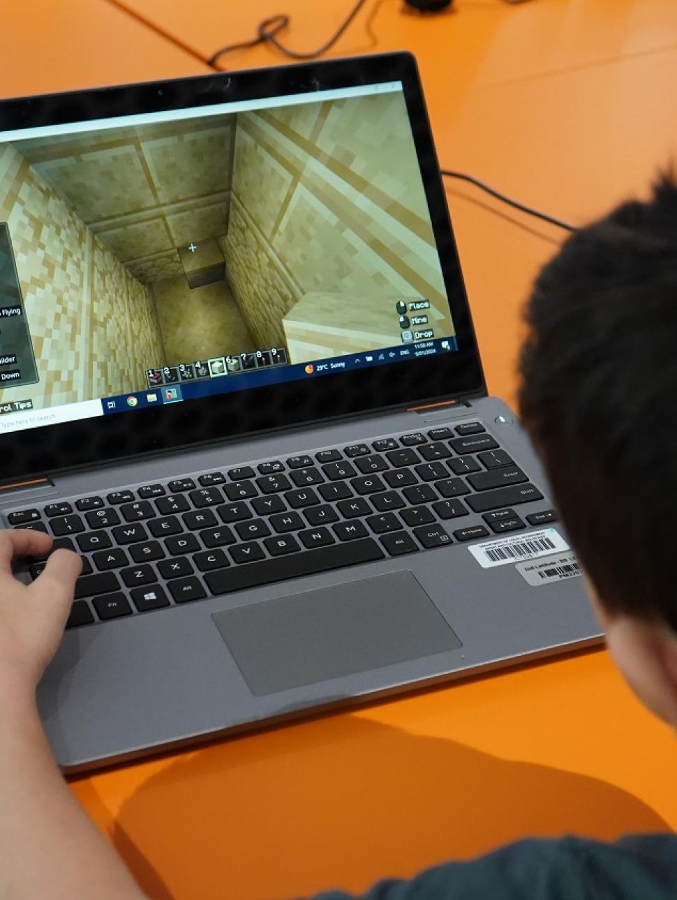 A young person sits at a bright orange table top and plays Minecraft on a silver and black laptop. The appear to be building a white limestone structure as per the last session's Egypt theme.