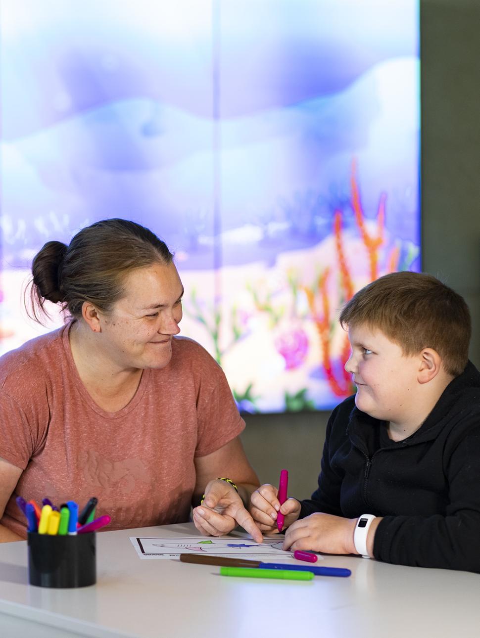 A parent with a child is sitting at a table colouring with a digital screen in the background depicting a sea scene