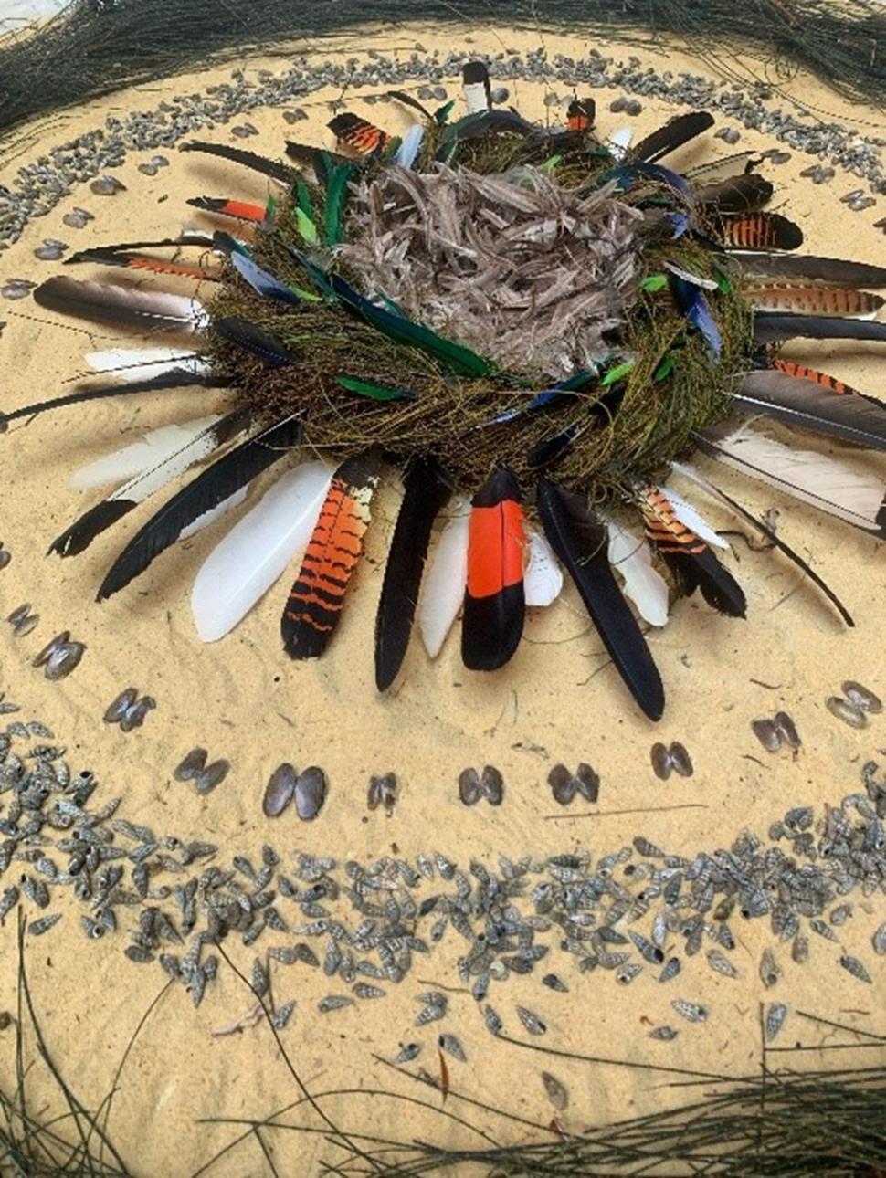 Circular artwork made from organic materials, including feathers, sand, plants and shells.  Centre piece of the work looks like a bird’s nest.  