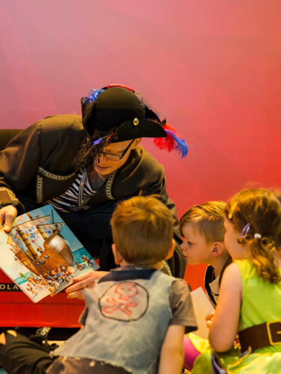 An adult dressed in a pirate costume holds a picture book and reads to a group of children who gather in front to look and listen