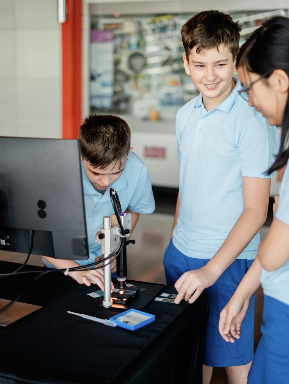 Three students in light blue polo shirts and dark blue shorts stand around a table with a black table cloth and a metal microscope on top. One child looks into the microscope while the two talk about the activity.