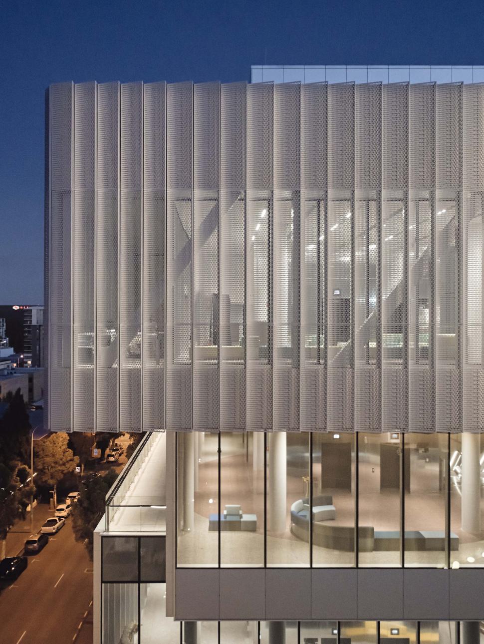 An exterior shot of the second and third levels of the WA Museum, peaking through the silver mesh detailing across the large windows into the brightly lit halls containing modern furnishings. The building is atmospherically lit against the deep blue of the Perth evening and yellow fo the nightly streetlamps
