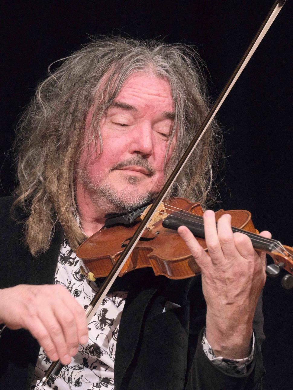 A half portrait image of musician Rupert Guenther, a white man with dark blonde/grey hair. He is holding a violin and is mid bow. He is wearing a black velvet jacket and a white and black patterned shirt.  