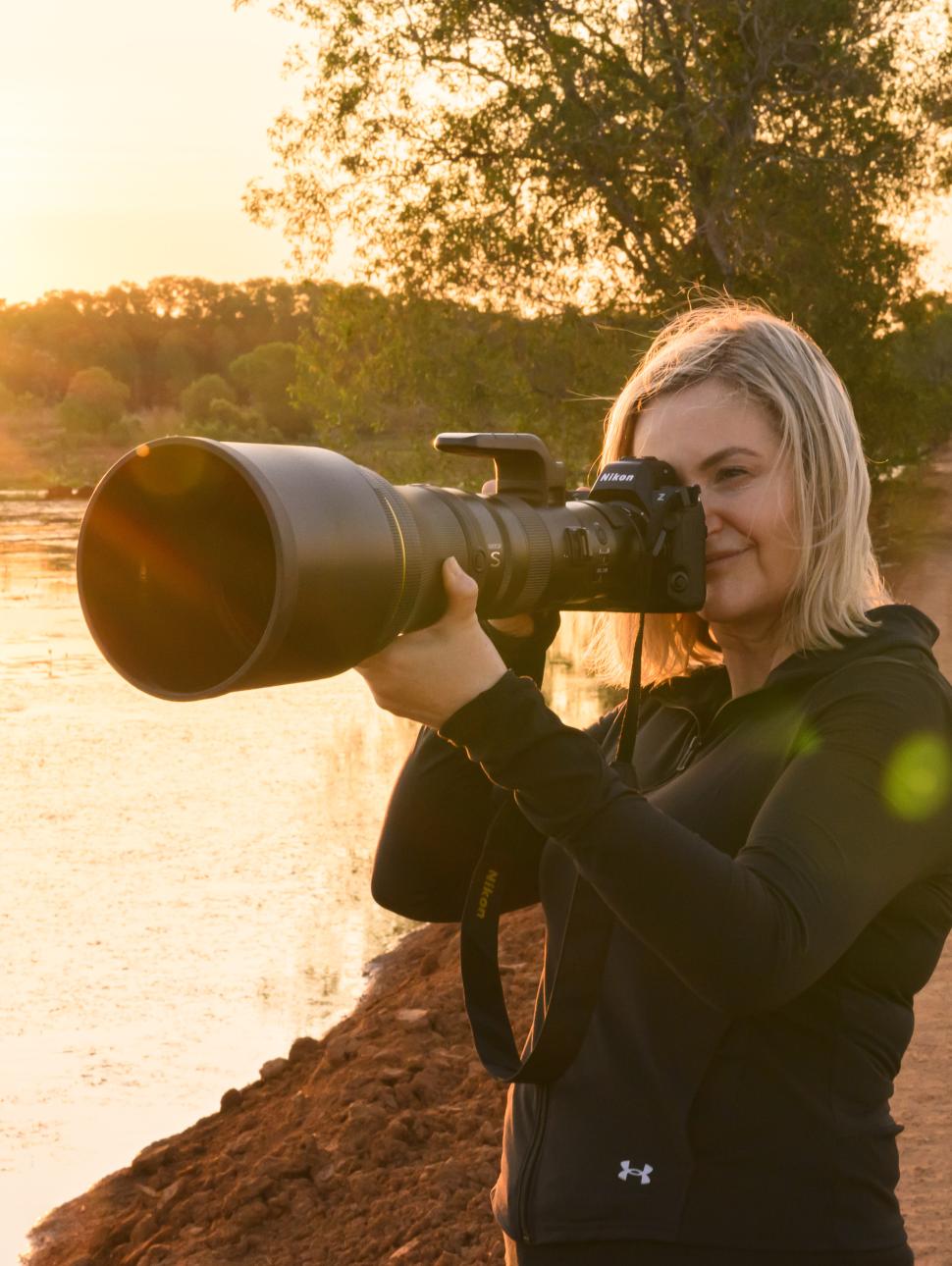 a person with blonde shoulder length hair holds a camera looking through it with a long lens. There is a sunset and lake in the background