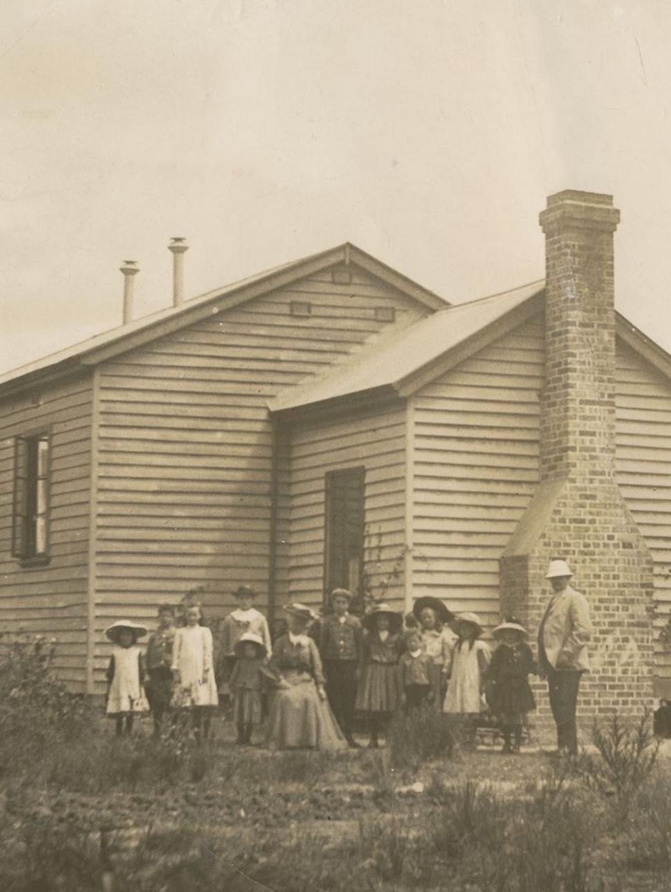 a historic sepia photograph of an old building hall with a group of people adults and children gathered in front of the building