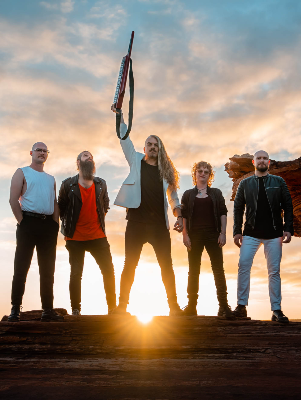 The band Voyager stand on a large flat rock in Kalbarri. The sun is rising behind them casting a red glow. Danny the lead singer is standing in the middle with the rest of the band either side of him. He is holding his keytar aloft