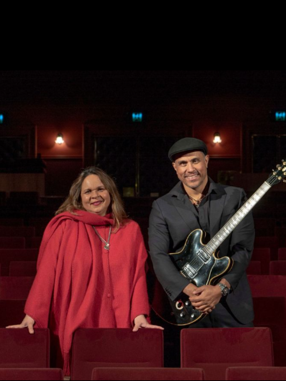Musician's Gina Williams and Guy Ghouse stand among red velvet theatre chairs. Gina is wearing a red coat and silver necklace and Guy is wearing a black suit and hat, he is also holding a black guitar. Both are looking at the camera and smiling.  