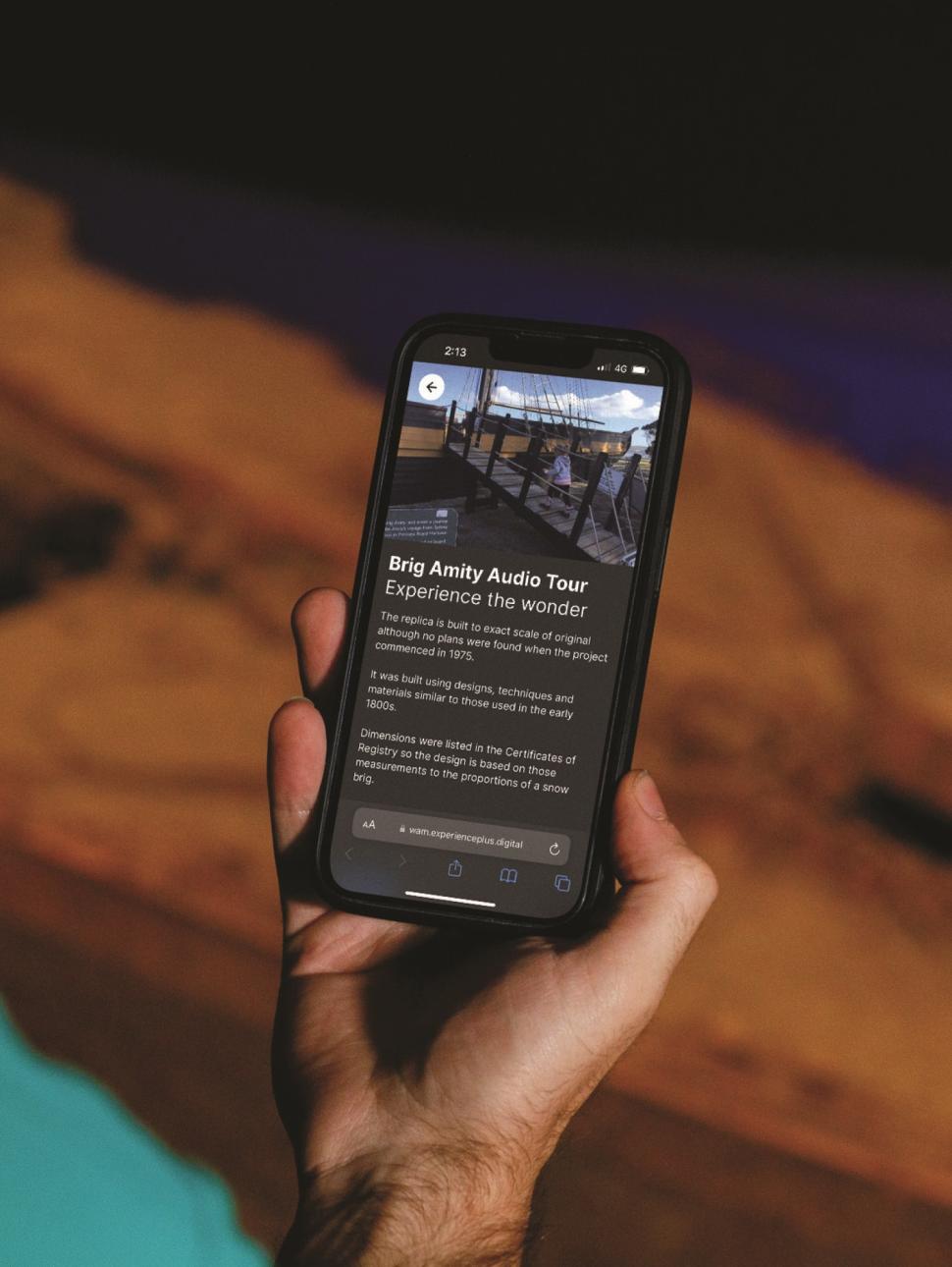 A hand holding a phone with the screen facing us. The screen shows the text Brig Amity audio tour with a picture of a walkway to walk to a ship. 