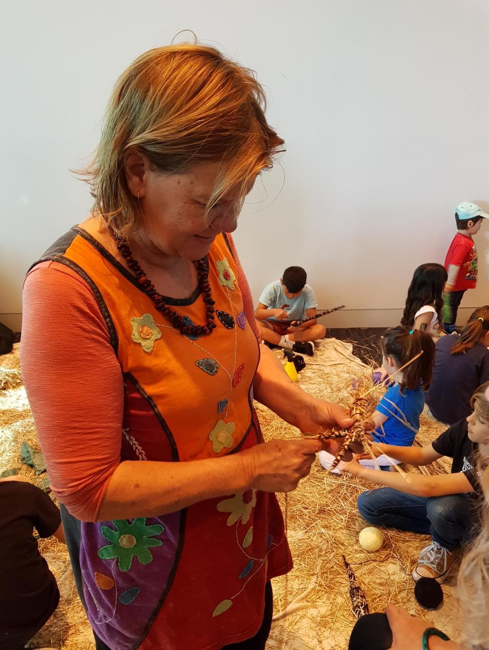 Woman standing weaving raffia with children sitting on floor behind her also weaving