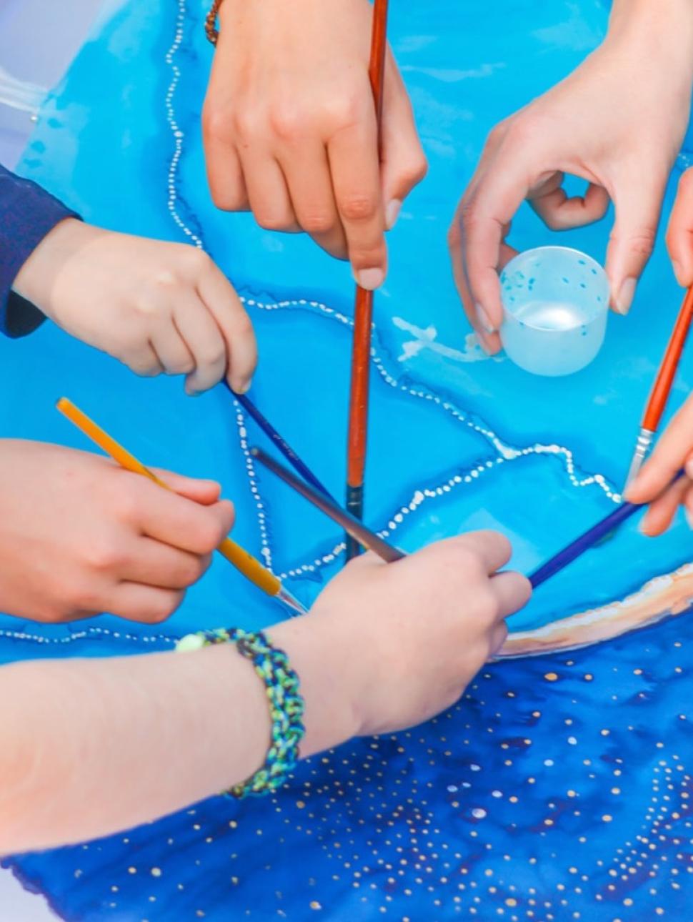Children's hands holding paintbrushes above a blue communal artwork 