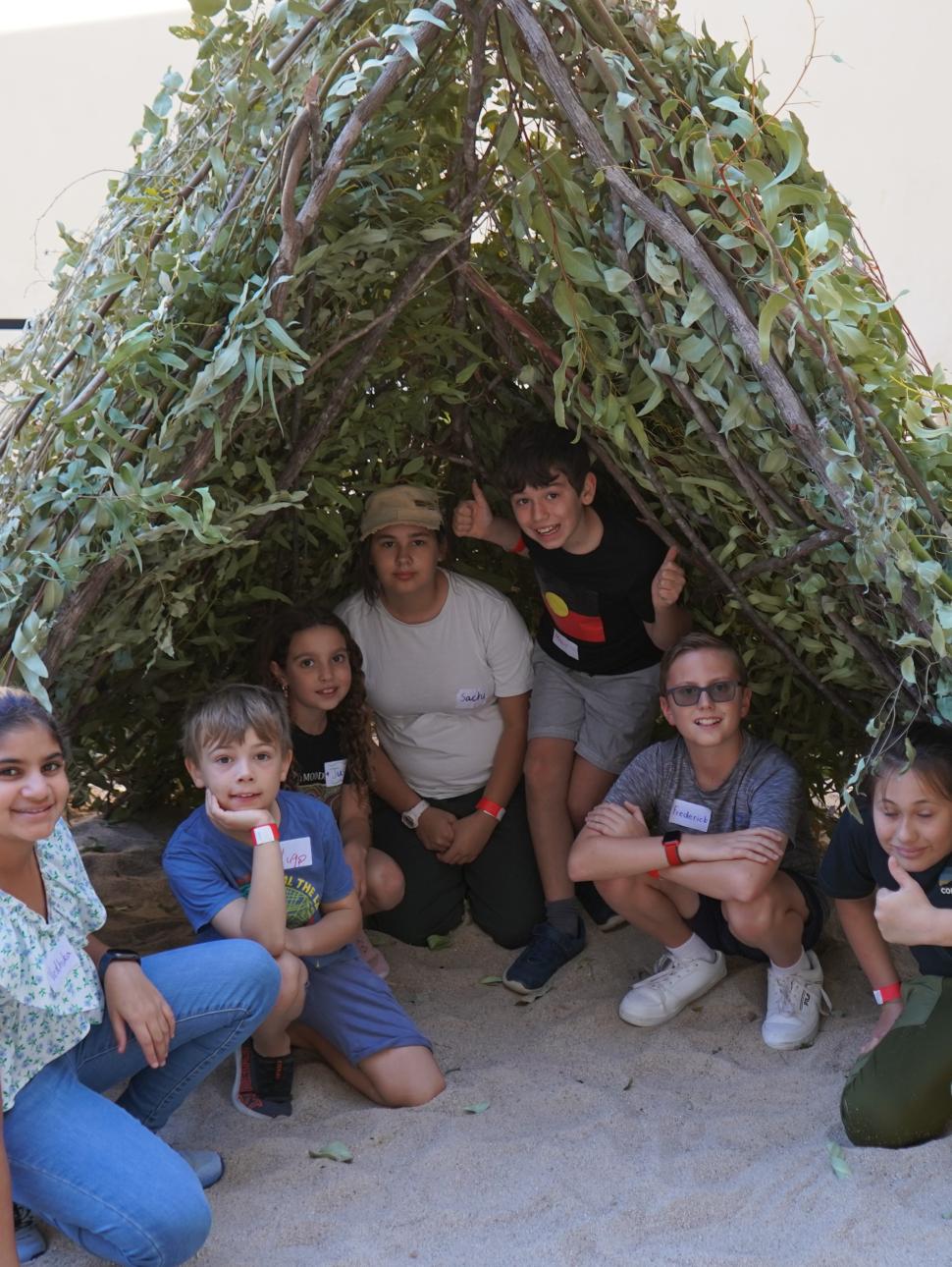 The image shows a small group of young people sitting inside a kalak, Nyoonger winter shelter, which they have collaborated to construct. The framework is made from strong brnaches and it is covered with leafy branches to keep the elements out. 