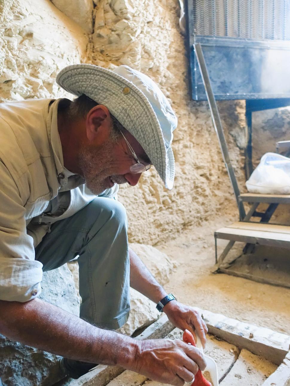 Man wearing hat and glasses crouched over ancient object brushing away dirt with paintbrush