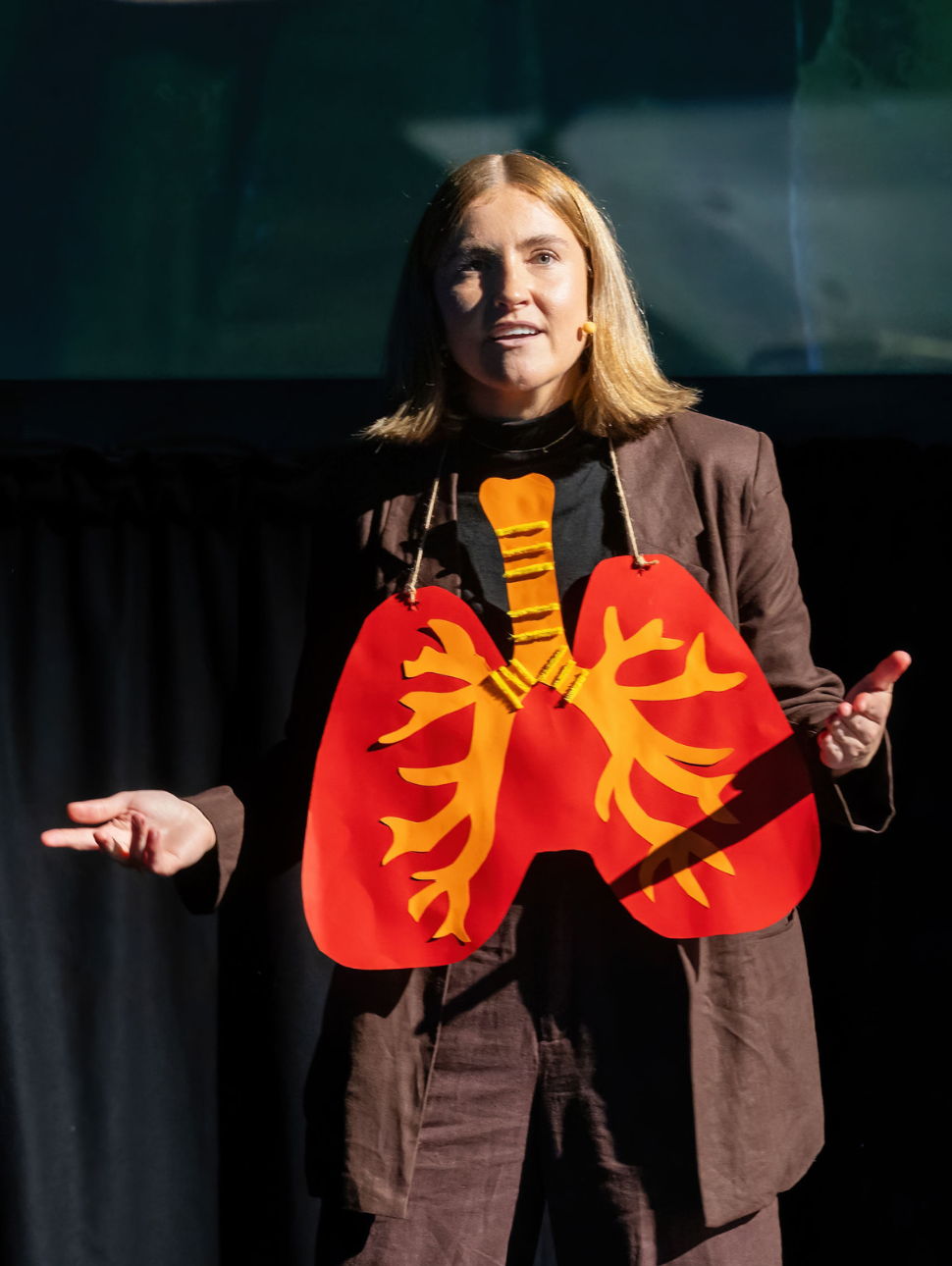 A blonde woman with cropped hair wears a paper heart around her neck, describing the anatomy 