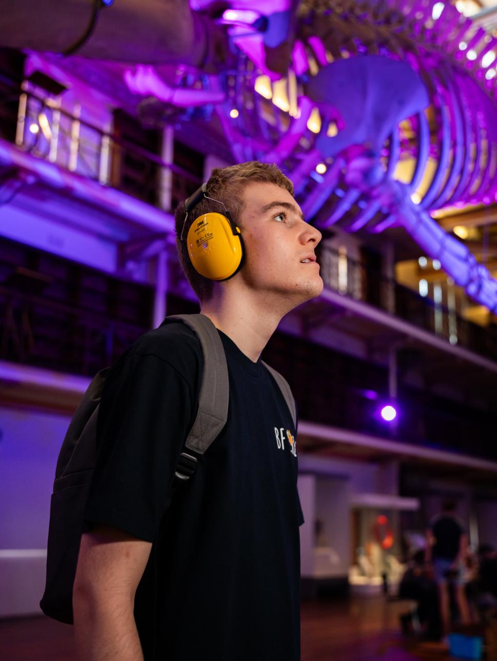 A young man wearing headphones stands in front of a whale skeleton, captivated by the majestic remains.