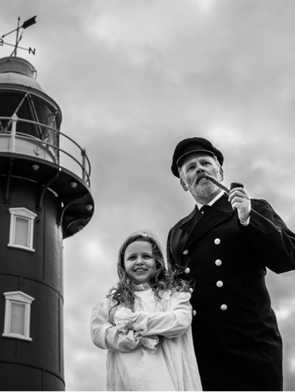 a black and white image of a lighthouse and two people standing in the foreground. One person is an adult, has a beard and wears a uniform smoking a pipe standing next to a young child with long hair and light coloured dress crossing their arms.