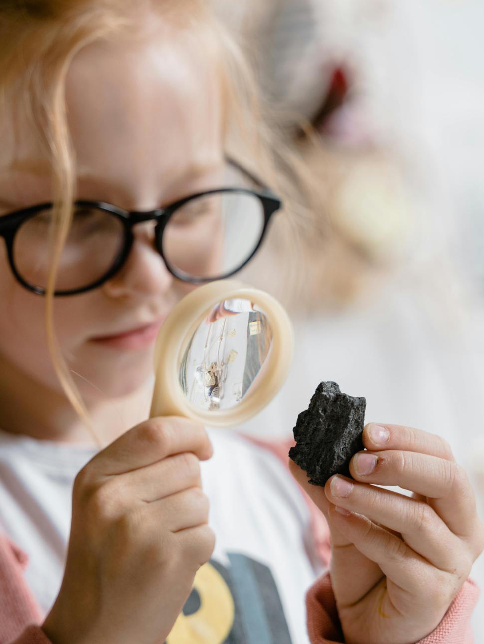 A yound child holds a dark coloured rock in one hand and holds a magnifying glass in another hand. They are looking at the rock in detail using the magnifying glass