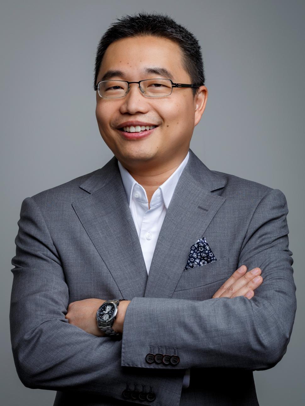 Marcus is an asian man with short black hair and glasses. He is wearing a grey suit and standing with his arms crossed smiling at the camera. 