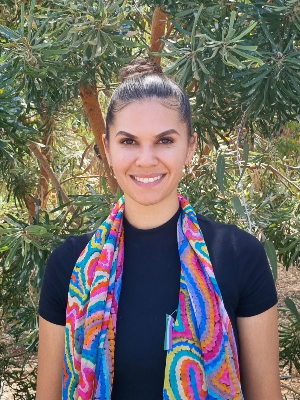 Rickeeta is a Nyoongar woman is standing against a native tree backdrop. She is wearing a black t-shirt and a bright coloured scarf and has her dark hair pulled back. She is looking at the camera and smiling.