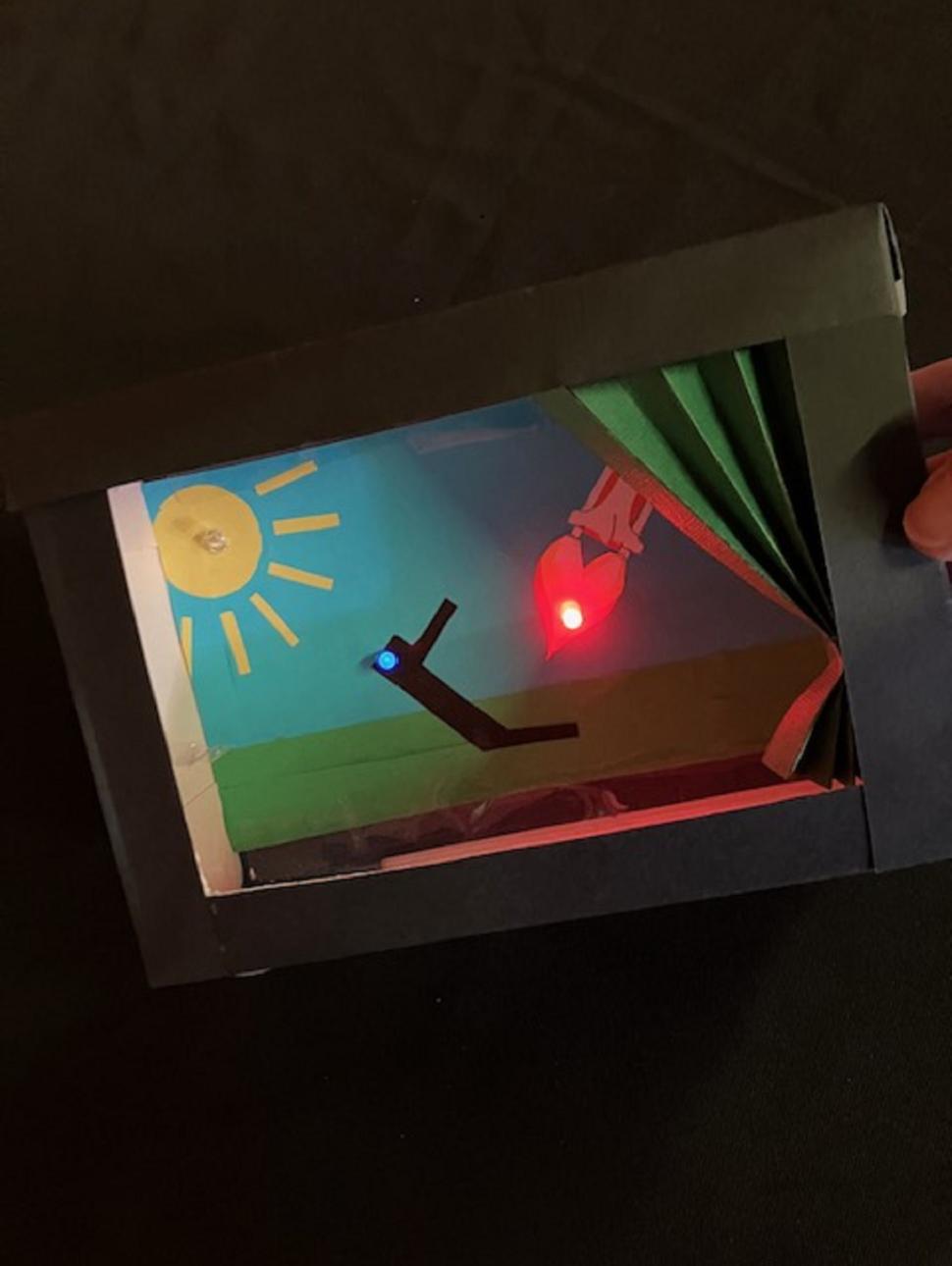 This image shows how a small cardboard box has been tunred in to a mini theatre using a backdrop, curtian and LED lights to highlight the stage. There is a yellow sun on the backdrop cut from yellow paper, also a red and blue LED light which illuminates.