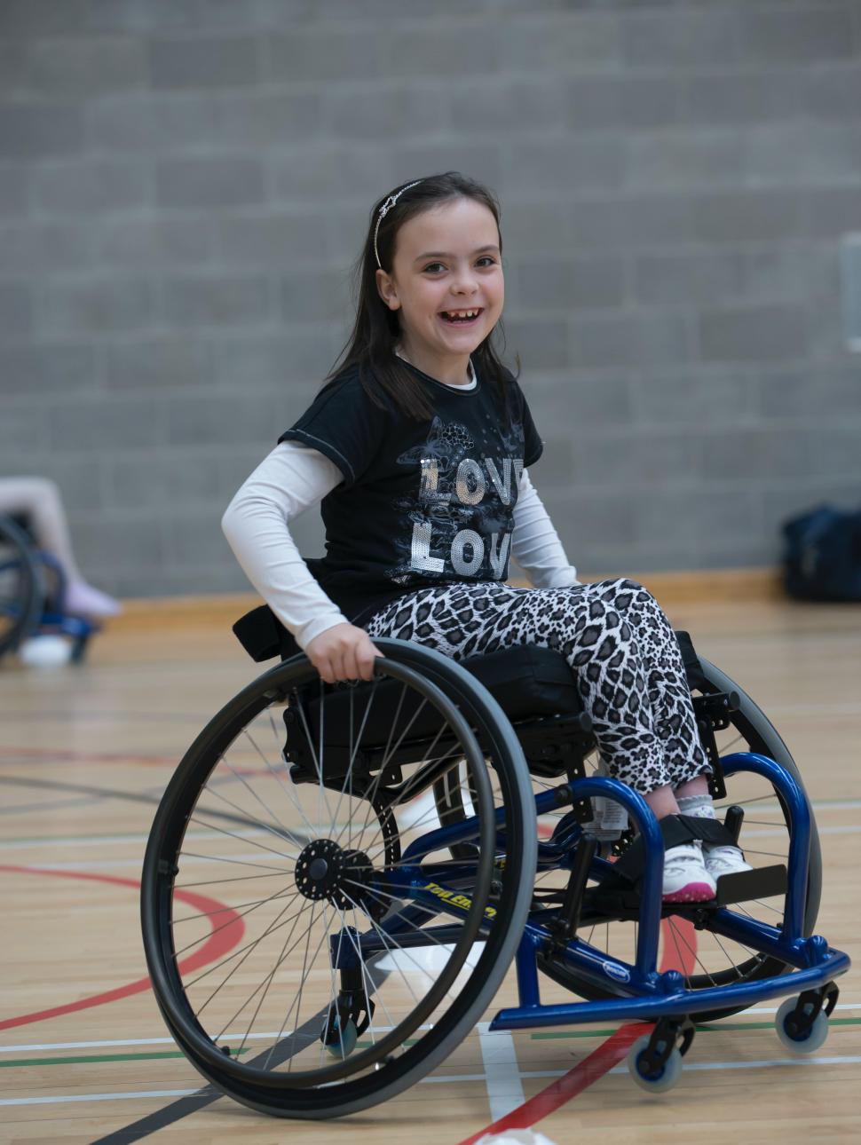 Girl in wheelchair and holding the wheels and smiling. Grey brick wall pictured in the background and a wooden floor with painted lines, indicating she is on a basketball court.