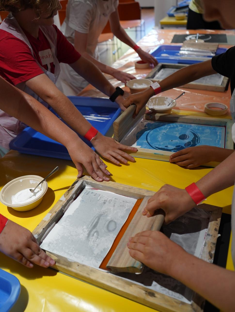 This image shows two chidren working together to make a screen print of the moon. They are using paint, a printing frame, sqeegie to spread the pain and paper on which to print the image. and 