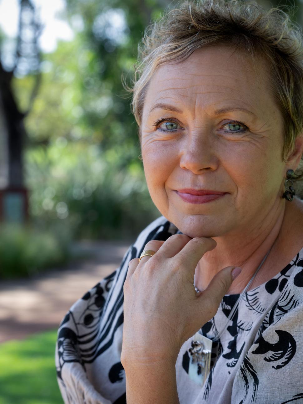 a close up of an adult looking at the camera leaning their chin on their hand. they are outside with trees and grass in the background