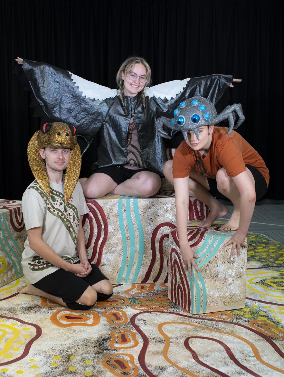 three young performers are dressed in colourful costumes of animals. they are standing on a decorated set with large block painted in cultural patterns. 