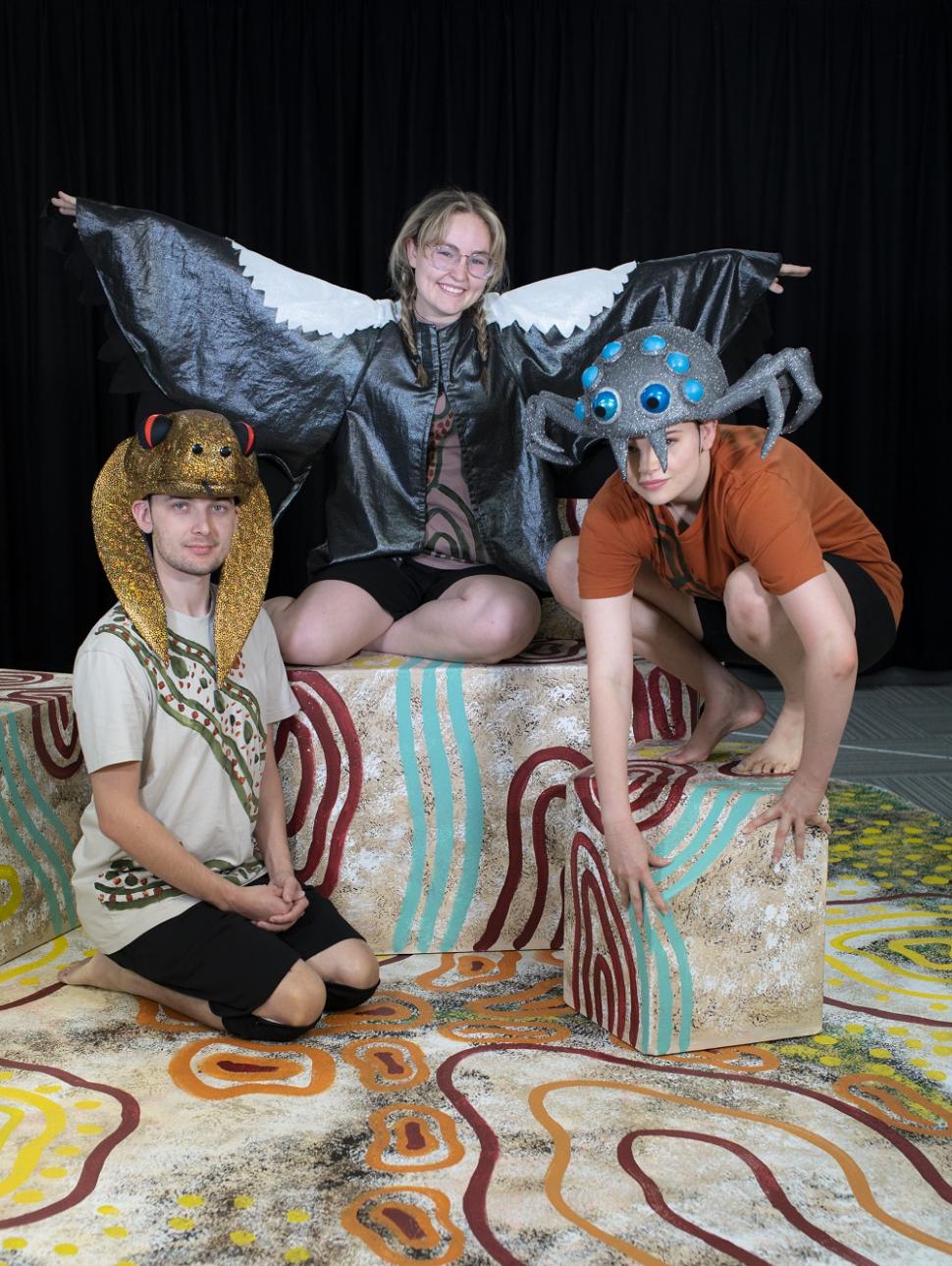 three young performers are dressed in colourful costumes of animals. they are standing on a decorated set with large block painted in cultural patterns.