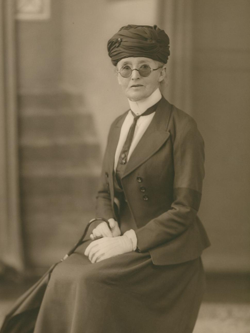 a woman dressed in posh clothing sits with hands crossed on her lap. The photograph is black and white.