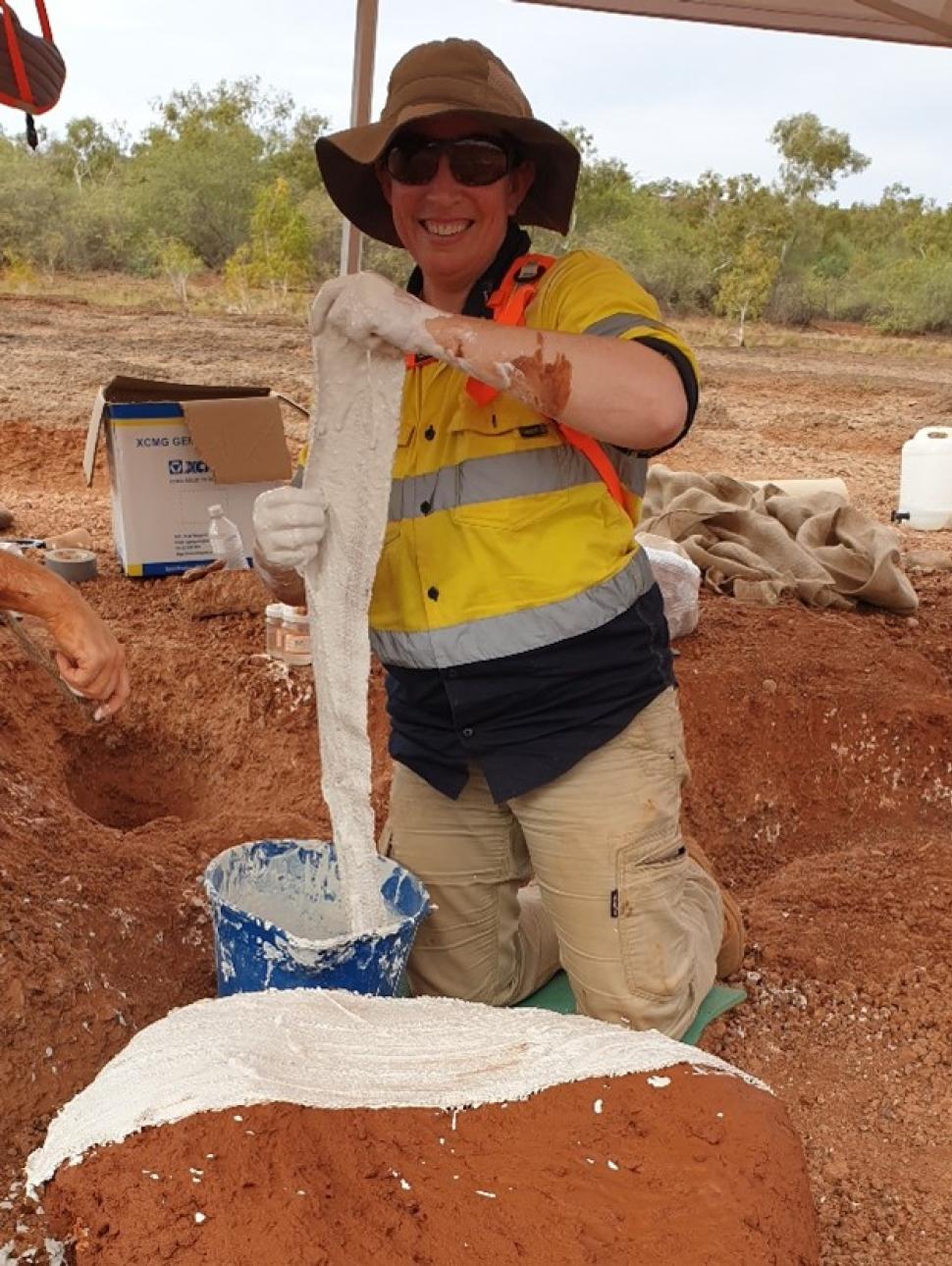 an adult person wearing high visability clothing, a hat and sunglasses is smiling as they kneel and hold up a long cloth wrap being taken out of a bucket of white liquid