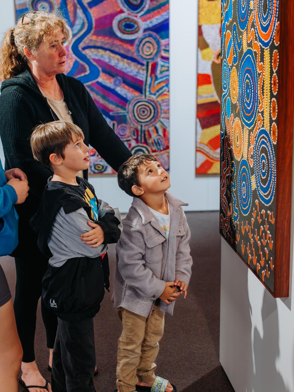 two oung children with their adults are looking at large artworks on display painted in traditional methods with vibrant colours