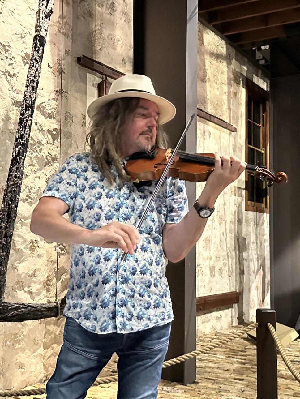 An adult person with a hat stands with a violin in a position to play the instrument In the background is an anchor and part of a shipwreck