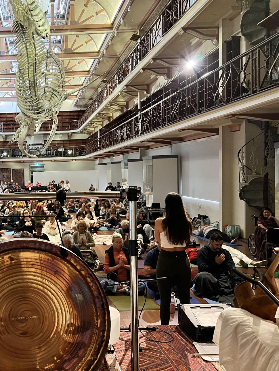A large gong takes place on a stage surrounded by musicians. An immersed audience faces the stage among the balcony's, stairs and exhibits of Hackett Hall.
