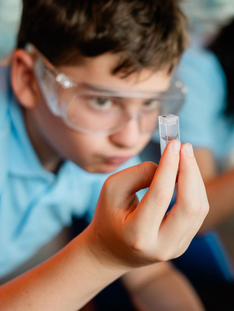 Boy wearing safety glasses inspects small scientific sample of marine life in a vial.