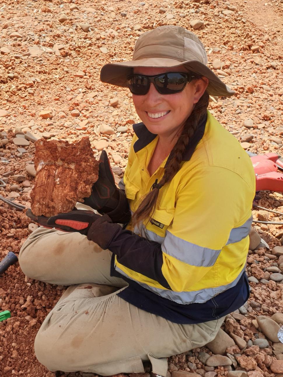 Lady wearing hat and sunglasses crouched, smiling at camera holding fossil