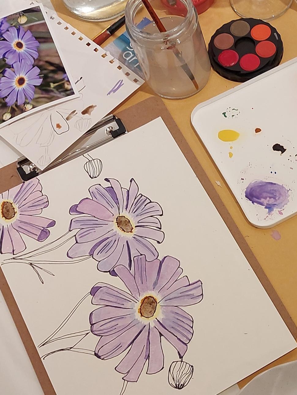 This image shows an example of a purple wildflower painted using watercolour paints. It also has some paints and a water jar with paintbrush next to it.