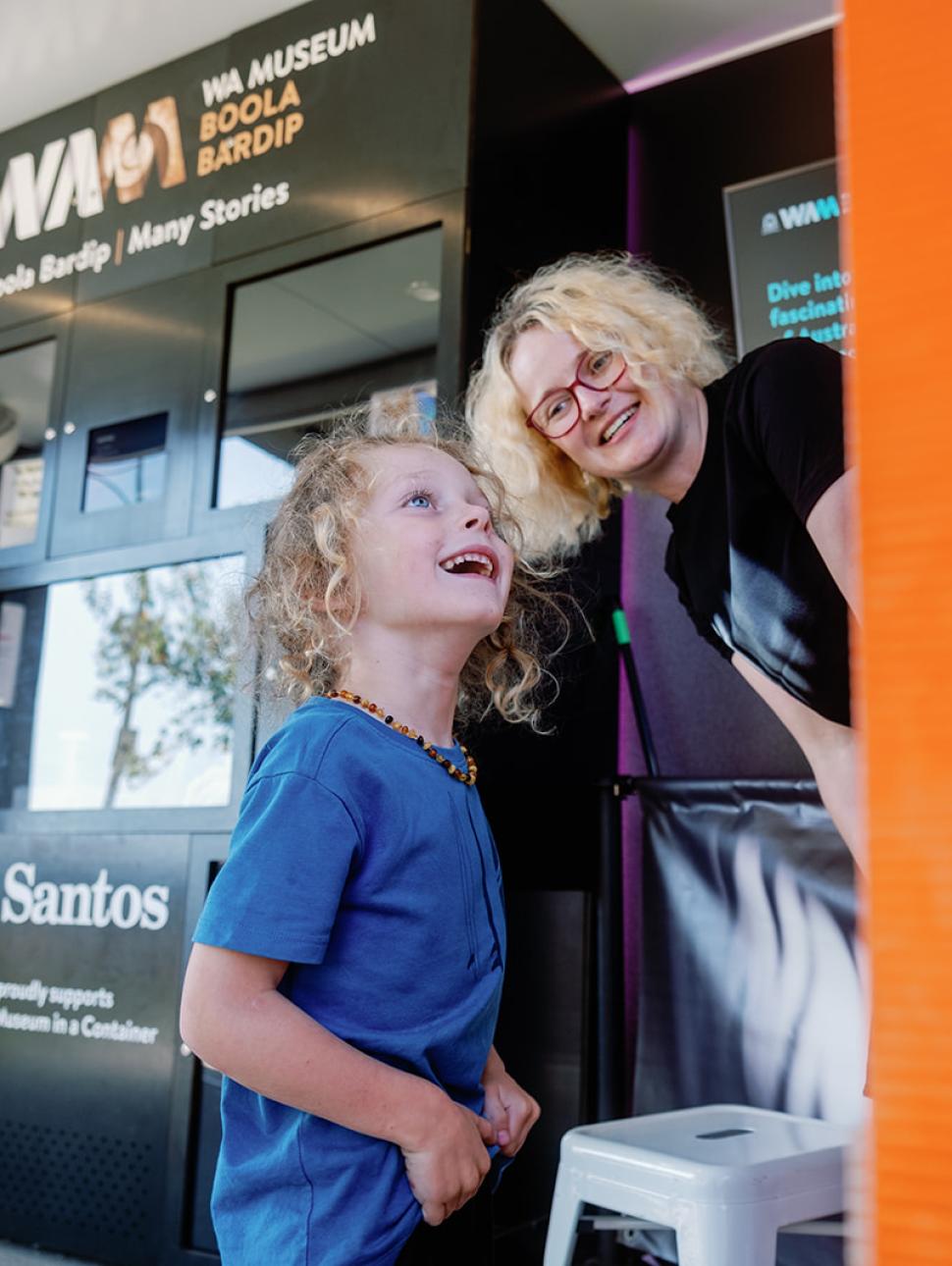 a young child looks up to a panel and smiles with an adult staff member in the background assisting