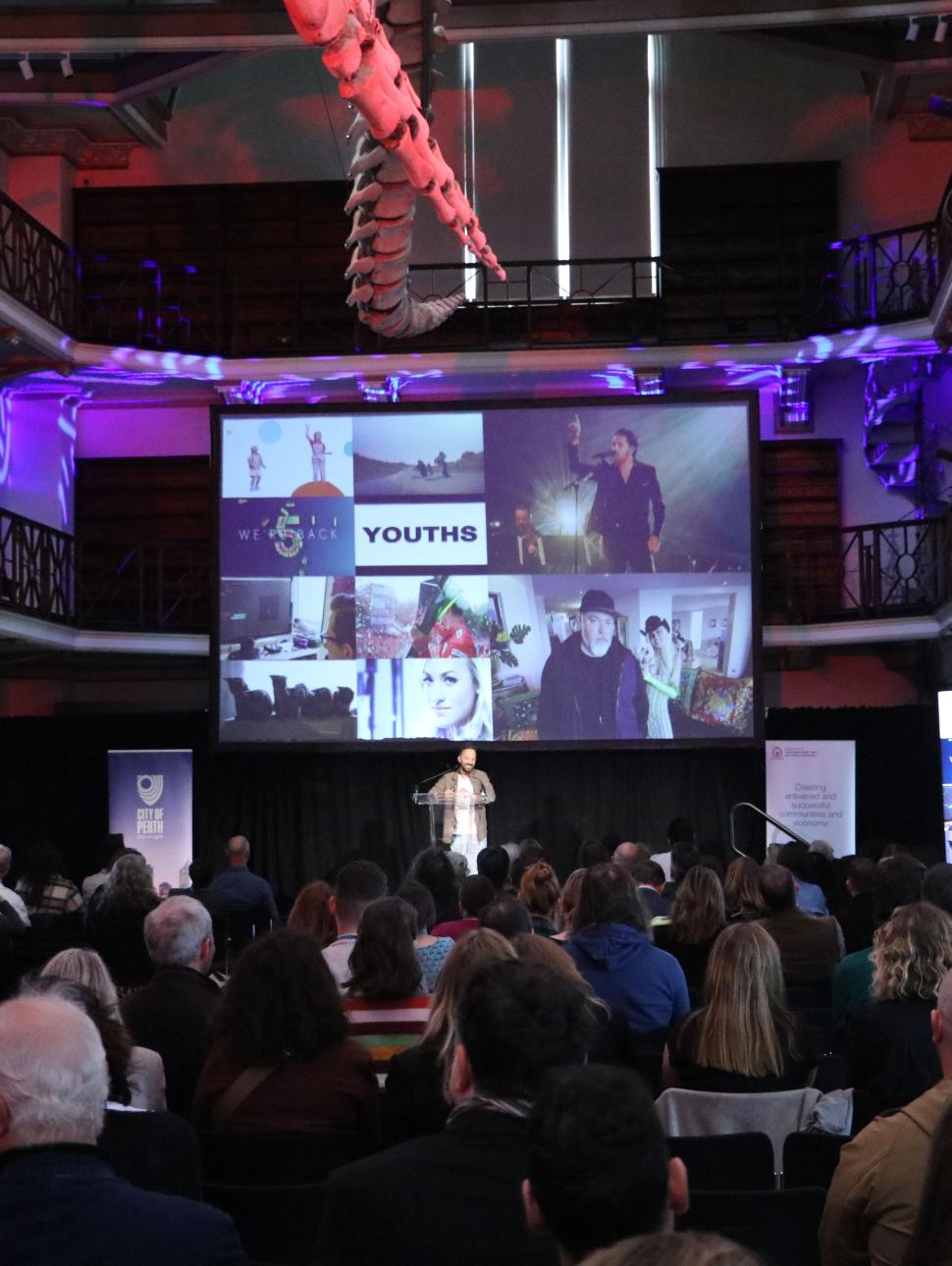 A large hall filled with people all facing a large screen and a male speaker at a lectern