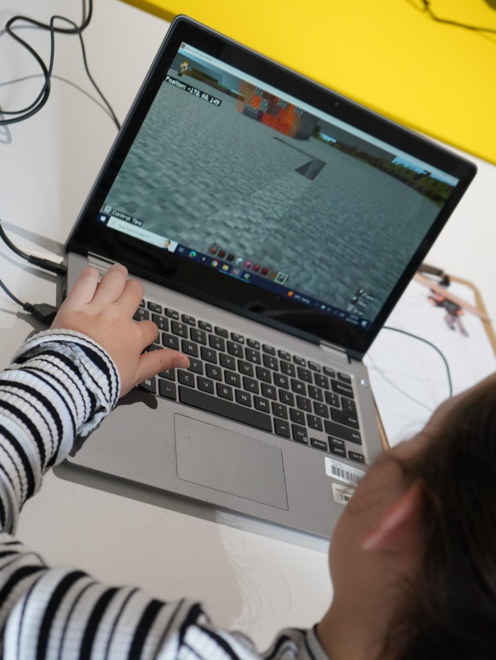 Looking over the shoulder of a young girl in a  striped T shirt. She is playing Minecraft on a laptop