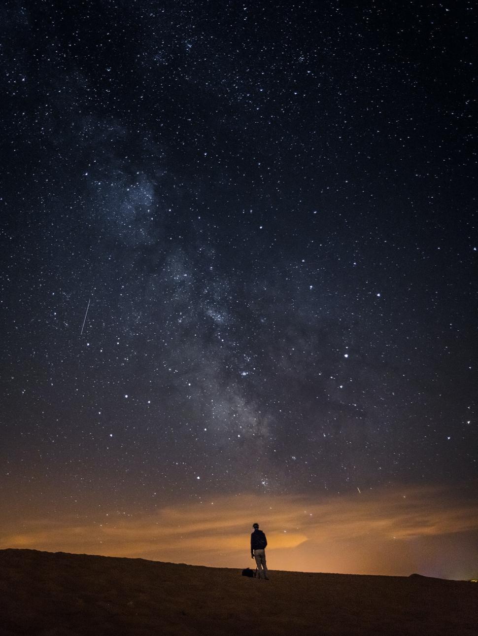 Image shows the night sky with many stars. The sun is in thwe final stages of settling on the horizon. A person is silhouetted in the distance. 