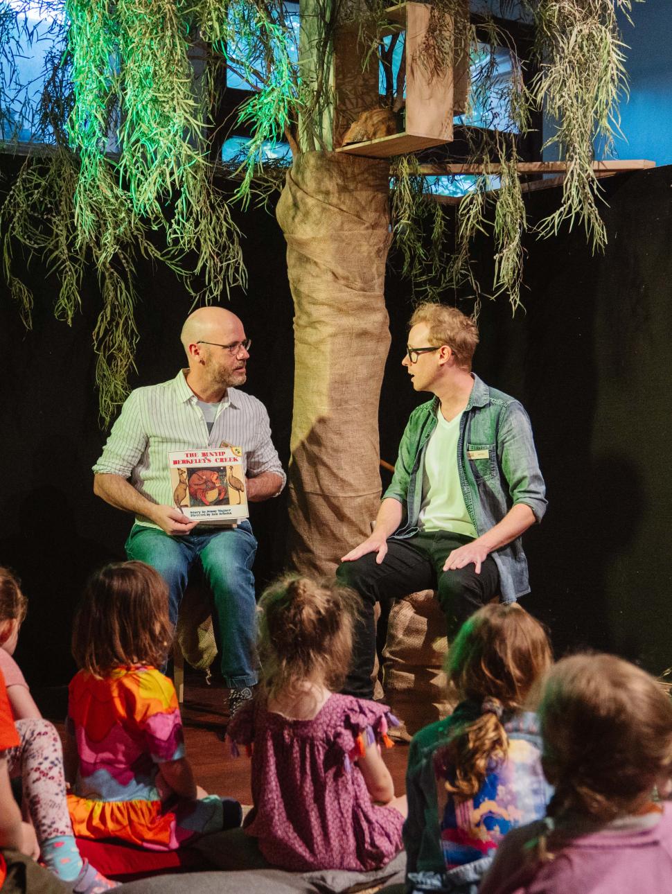 two adults on astage sit and read a story to a crowd of young children